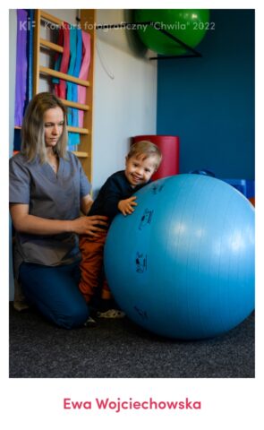 Zdjęcie przedstawia pracę fizjoterapeutki z dzieckiem z Zespołem Downa. Jest to jeden z najbardziej znanych i
powszechnych wrodzonych zespołów genetycznych. Leczenie dzieci jest złożonym procesem, który powinien
uwzględniać współpracę wielu specjalistów w tym również pracę fizjoterapeutów. Program rehabilitacji ma kluczowe
znaczenie w rozwoju dzieci z zespołem Downa. Wczesna i wieloprofilowa rehabilitacja dzieci umożliwia poprawę
funkcji somatomotorycznych oraz osiągnięcie optymalnego poziomu rozwoju. Chłopiec jest chętny do nawiązywania
nowych znajomości i dobrze reagują na bliskość drugiego człowieka – fizjoterapeutki. Fizjoterapeutka podczas zajęć
ruchowych stale śledzi zmiany nastroju oraz reakcje emocjonalne chłopca, a także kładzie duży nacisk na wzajemną
komunikację. Ćwiczenia równoważne, zwiększające koordynację oraz poprawiające znajomość schematu własnego
ciała - w tym celu zastosowano ćwiczenie z piłką. Mały pacjent zdążył dobrze poznać fizjoterapeutkę, dlatego czuje się
przy niej bezpiecznie i chętnie wykonuje wszystkie ćwiczenia.